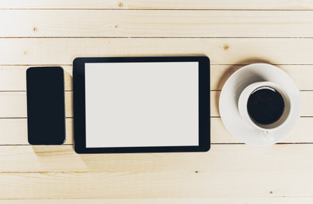 Black smartphone and tablet with empty screen beside coffee cup on wooden table