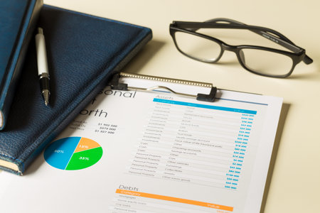 Calculating personal finances with a balance sheet, notebooks, pen, and glasses on a desk