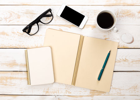Desk setup with notebooks, smartphone, coffee cup, and glasses on wooden table