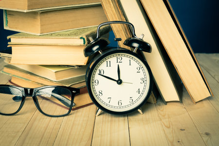 Cluttered desk with books, glasses, and alarm clock shows the essence of study timeの写真素材
