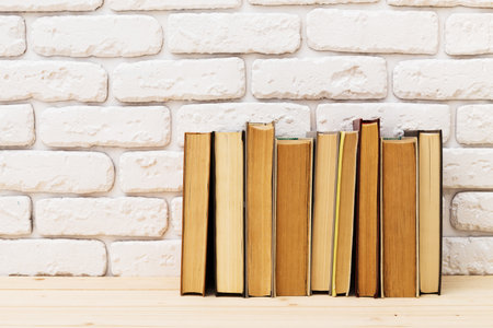 Rows of vintage books arranged on a wooden shelf against a white brick wallの写真素材