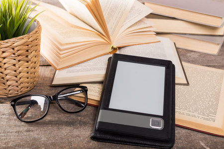 Books and an e-reader on a wooden table with glasses and a plant