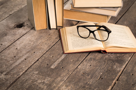 Reading glasses placed on an open book next to a stack of books on a wooden tableの写真素材