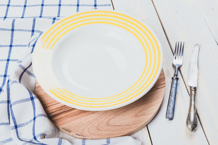 Empty plate with yellow stripes on wooden board next to fork and knife on tableclothの写真素材