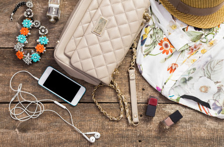 A collection of fashion accessories, a smartphone, and cosmetics arranged on a wooden table