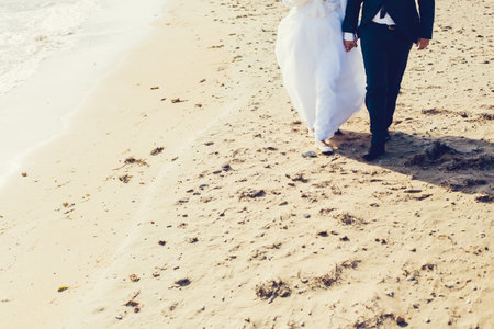 Couple walking hand in hand on sandy beach during romantic wedding ceremony in sunlightの写真素材