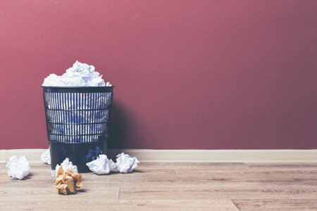 Waste basket filled with crumpled paper near a red wall and wooden floor in an indoor space