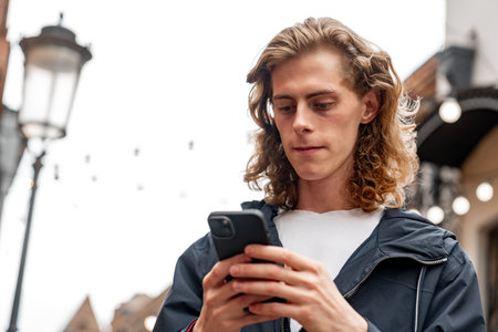 Young man checks phone while walking through urban street in cool weather with soft lightingの写真素材