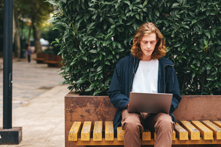 Young man focused on laptop while sitting on bench in urban park settingの写真素材