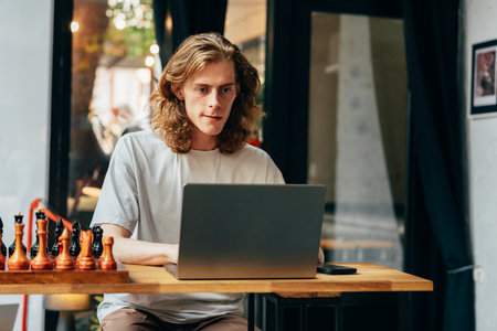 Young man working on laptop at a cafe with chess pieces nearby during the dayの写真素材