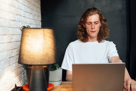 Young man working on laptop at a cozy desk with warm lighting in a modern spaceの写真素材