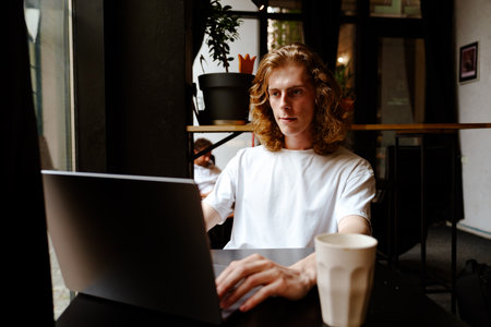 Young man working on laptop in a cozy cafe with a warm ambianceの写真素材