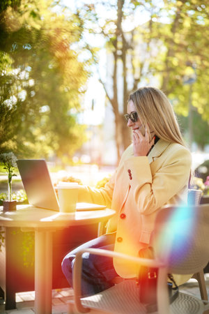 Working on a laptop outdoors while talking on a phone in a sunny cafe settingの写真素材