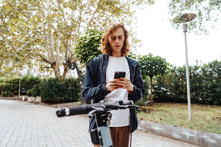 Young man checking phone while standing near electric scooter in urban park settingの写真素材