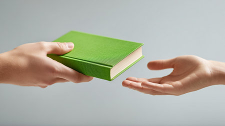 Person handing over a green book to another individual with an outstretched handの素材