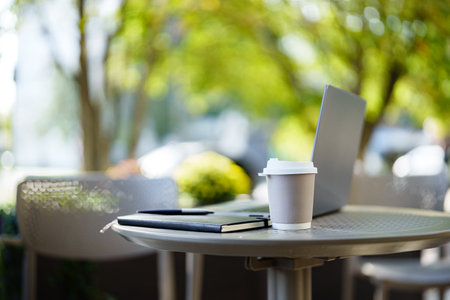 Laptop and coffee cup on table outdoors in a sunny urban settingの写真素材