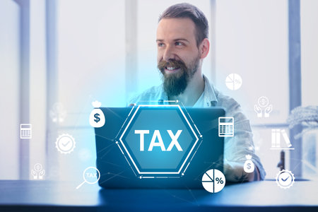 Man smiling while working on laptop with tax graphics and icons in a modern office settingの写真素材