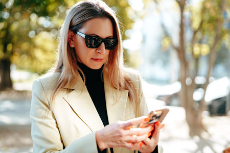 Woman in sunglasses checks her phone while walking in a sunny urban parkの写真素材