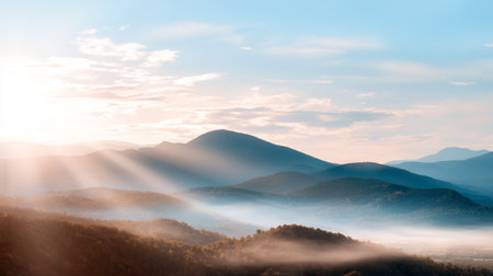 Mountain landscape under a clear blue sky with sunlight breaking through clouds at dawnの素材