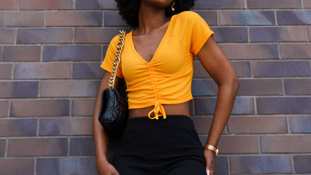 Young woman stands against a brick wall wearing a bright top and black pants while holding a bag