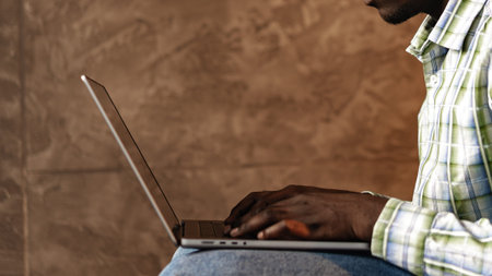 Man typing on a laptop while sitting against a textured wall in a casual settingの写真素材