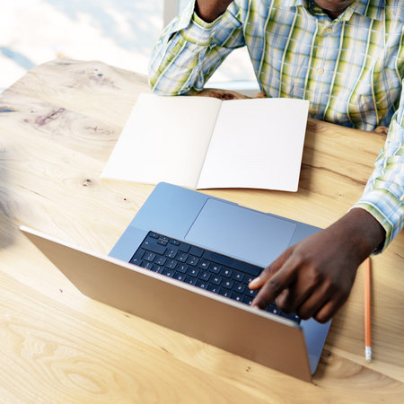 Person working at a laptop with an empty notebook on a wooden table in a bright setting