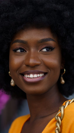 Woman with curly hair smiles while wearing yellow top and earrings, in a busy outdoor setting