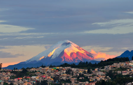 The majestic Cotopaxi volcano, with its snow-capped summit and bathed in the warm tones of a sunset, towers over the vibrant city of Quito, Ecuador. The colorful facades of the buildings in the foreground contrast with the imposing presence of the volcano in the background, highlighting the coexistence between the natural beauty of the Andes and urban life.の写真素材