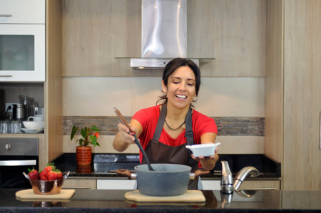 A Latin American woman of mixed race, with a happy and satisfied expression, pours a portion of food from a large pot into a small bowl in a well-equipped contemporary kitchen.の写真素材