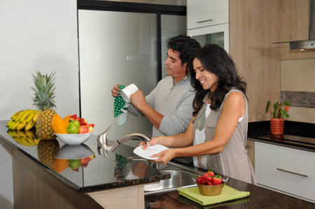 A happy Latin American couple collaborates on household chores, washing and drying dishes in a bright, modern kitchen, symbolizing teamwork and domestic harmony.の写真素材