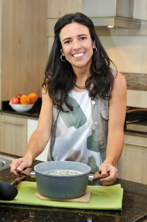 A middle-aged Latina woman smiles at the camera while holding a pot of freshly made soup in a modern kitchen. The image radiates a sense of well-being, home cooking, and joy.の写真素材