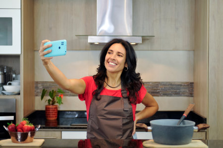 A middle-aged woman of Latin ethnicity smiles as she takes a selfie with her mobile phone in a modern kitchen. She is wearing an apron and appears to be enjoying the moment.の写真素材