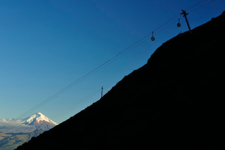 A stunning view of a cable car ascending a steep mountainside in silhouette, with the iconic snow-capped summit of Cotopaxi Volcano emerging under a pristine blue sky on the Ecuadorian horizon. The scene combines modern engineering with the grandeur of Andean nature.の写真素材