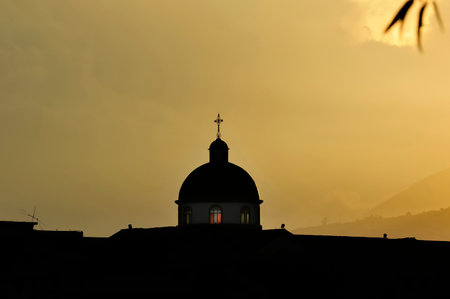 Silhouette of a church dome with a cross on top, standing out against a golden evening sky, with illuminated windows and a mountainous landscape in the background, evoking spirituality and tranquility.の写真素材