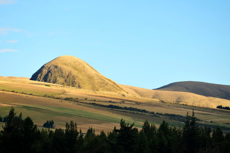 This image captures the serene beauty of a natural elevation overlooking a vast Andean landscape, bathed in the warm golden light of sunset. Cultivated fields and mountain formations in the distance stretch out under a clear sky, creating an atmosphere of tranquility and natural majesty.の写真素材