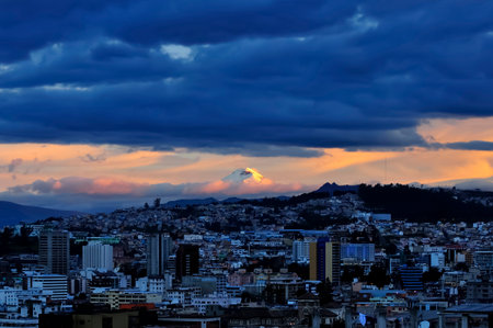 A curious panoramic view of the city of Quito at sunset, where the majesty of the Andes can be appreciated, with the Cotopaxi volcano framed by clouds and the city skyline.の写真素材