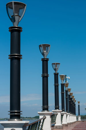 A row of sleek, modern streetlights stretches along a seawall under a vibrant, clear blue sky. The diagonal perspective creates a sense of depth and continuity.の写真素材