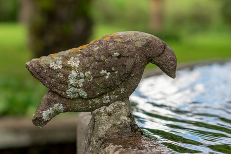 Close-up of an antique stone sculpture shaped like a bird or dove, covered in moss and lichen,...