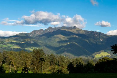 Pasochoa Volcano in the Ecuadorian Andes. A lush mountain landscape with green hills and dense forest, under a blue sky with white clouds and a visible moon.の写真素材