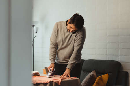 Latin man with braces ironing women's clothes in living room. Standing holding an outfit and smiling. Concept of equal chores at home.の写真素材
