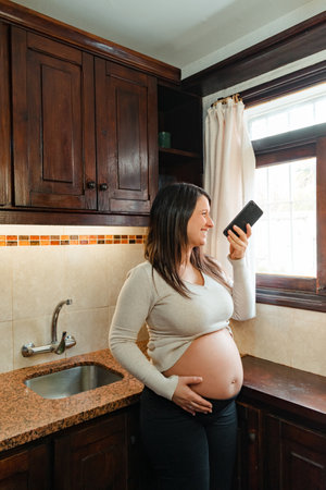 pregnant Hispanic woman, recording audio on her cell phone, while holding her belly and looking out the kitchen window.の写真素材