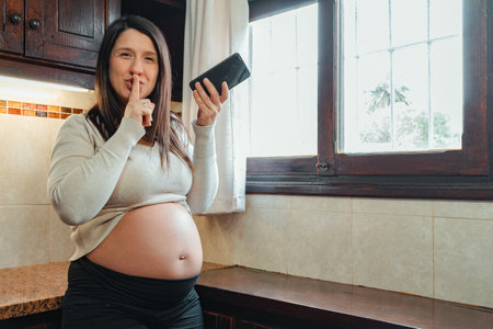 pregnant Hispanic woman, recording audio on her cell phone, while holding her belly and looking out the kitchen window.の写真素材
