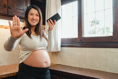 pregnant Hispanic woman, recording audio on her cell phone, while holding her belly and looking out the kitchen window.の写真素材