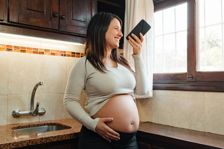 pregnant Hispanic woman, recording audio on her cell phone, while holding her belly and looking out the kitchen window.の写真素材