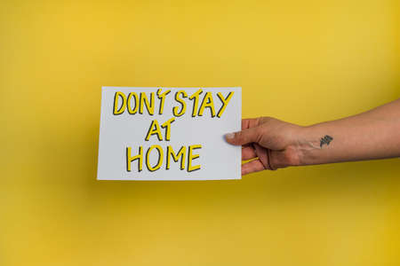 A closeup shot of a woman holding a paper with the word "DON'T STAY AT HOME" on a yellow backgroundの写真素材