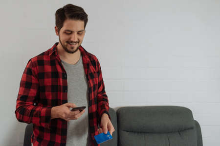 Latin man in red plaid shirt and beard making an online purchase indoors.の写真素材