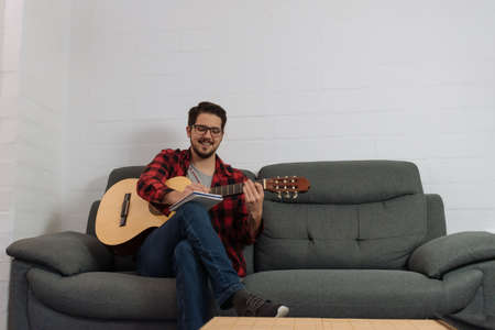 Latin hipster in glasses and red plaid shirt writing his musical composition in a notebook while holding the guitar.の写真素材