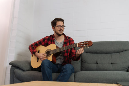 Latin hipster in glasses and red plaid shirt tuning the guitar on the couch of a house.の写真素材