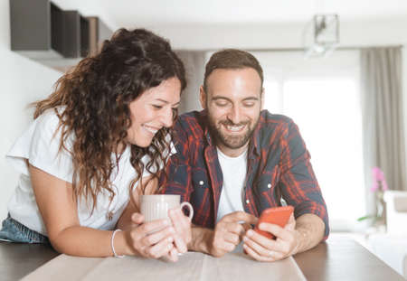 Young couple laughs while using smartphone - couple in love having breakfast together and reading the latest news using mobile phoneの写真素材