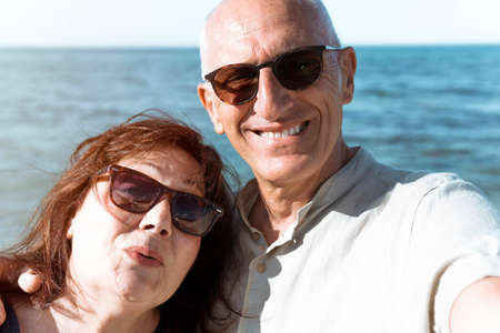 Middle-aged couple takes a selfie on the beach during a vacation - Happy elderly couple having fun in their free time - Active retirees having fun traveling the world concept - flare on backgroundの写真素材